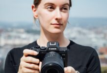 Canon EOS R8 Mark II : le plein format compact gagne la stabilisation Une photographe tient le Canon EOS R8 Mark II équipé d'un zoom RF devant un paysage urbain flou