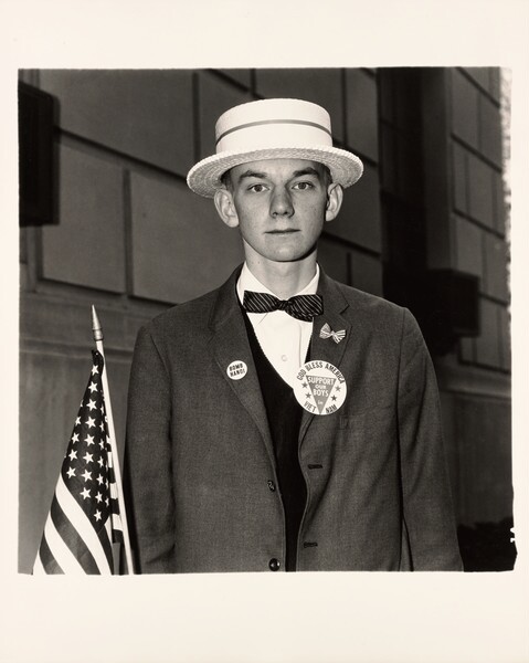 Jeune homme en canotier, nœud papillon et badges, à côté d’un drapeau américain, photographie de Diane Arbus (1967)