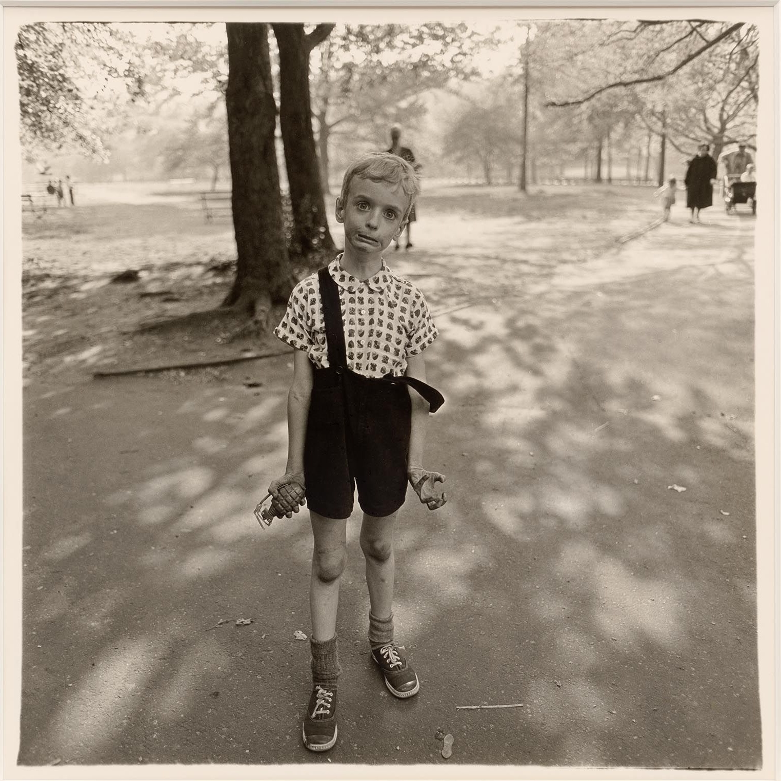 Garçon maigre en short à bretelles tenant une grenade en plastique et grimaçant dans Central Park, photographie de Diane Arbus (1962)