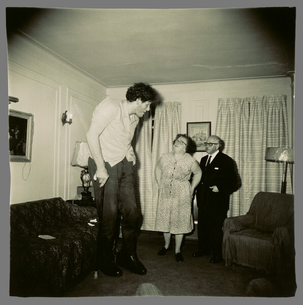 Eddie Carmel, homme de très grande taille, voûté dans le salon familial face à ses parents, photographie de Diane Arbus (1970)