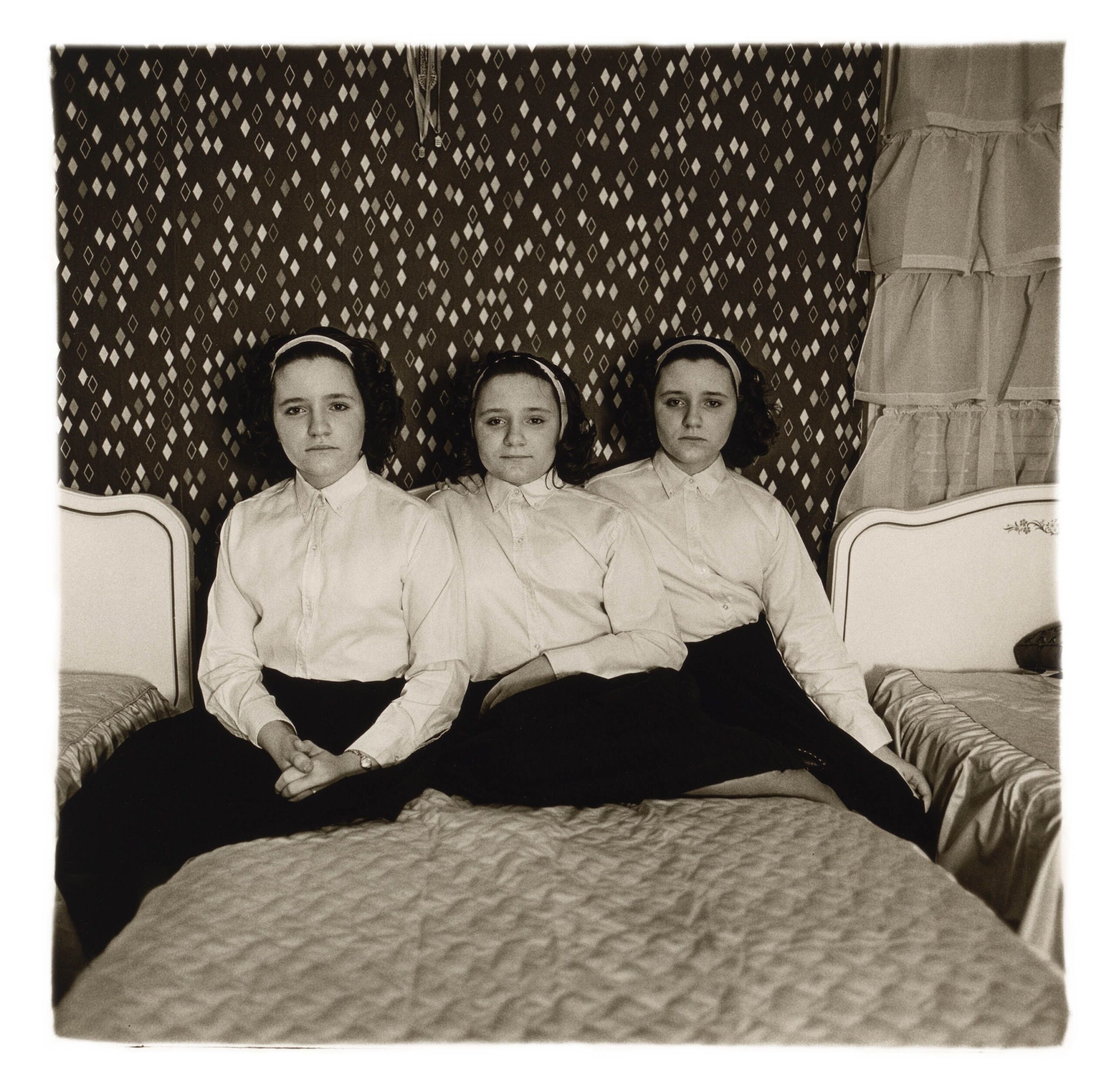 Trois sœurs triplées en chemisier blanc assises sur un lit devant un papier peint à pois, photographie de Diane Arbus (1963)