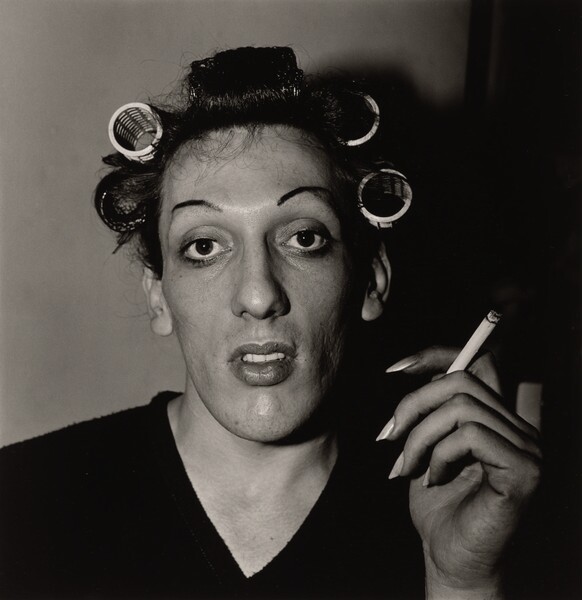 Jeune homme maquillé aux sourcils épilés, bigoudis dans les cheveux et cigarette à la main, photographie de Diane Arbus (1966)