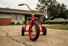 William Eggleston, l’homme qui a fait entrer la photographie couleur au musée Tricycle rouge photographié au ras du sol sur une allée en béton fissuré, devant une maison de banlieue américaine et une voiture ancienne